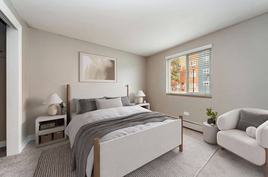 A modern bedroom at Esprit Cherry Creek features a double bed, two nightstands with lamps, a cushioned armchair, a window with blinds, and neutral-toned decor.