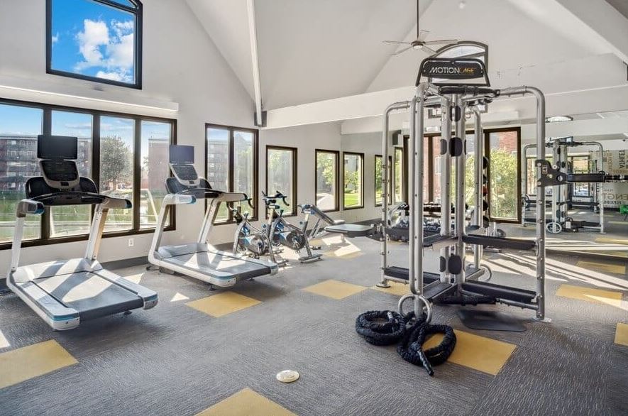 Bright gym room at Esprit Cherry Creek with treadmills, stationary bikes, a cable machine, battle ropes, large windows, and gray carpet with yellow accents.
